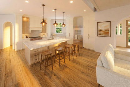 A bright, modern kitchen with a large central island, wooden bar stools, wood flooring, and warm white cabinetry. A white upholstered chair is visible in the foreground, and the room is lit by recessed ceiling lights and pendant lights.