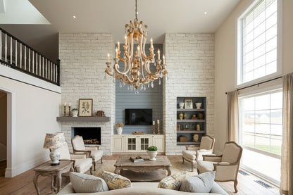 A large, elegant multi-light chandelier with gold-toned metal and clear crystal accents hangs in a bright, modern living room with a white brick fireplace and high windows.