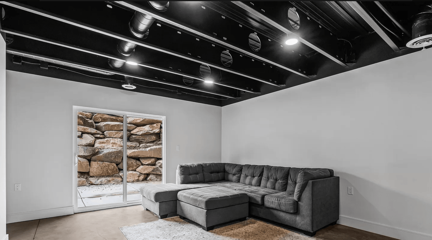 Modern No.10 house plan basement apartment with exposed black ceiling beams, gray sectional sofa, and sliding glass door to rock retaining wall.